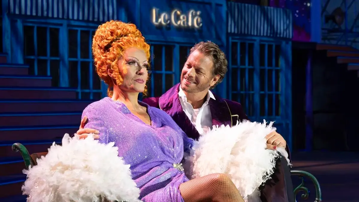 Eine Frau mit orangefarbenen Locken und einem lila Kleid sitzt auf dem Schoß eines Mannes vor einem blauen Gebäude mit dem Schild 'Le Cafe'.