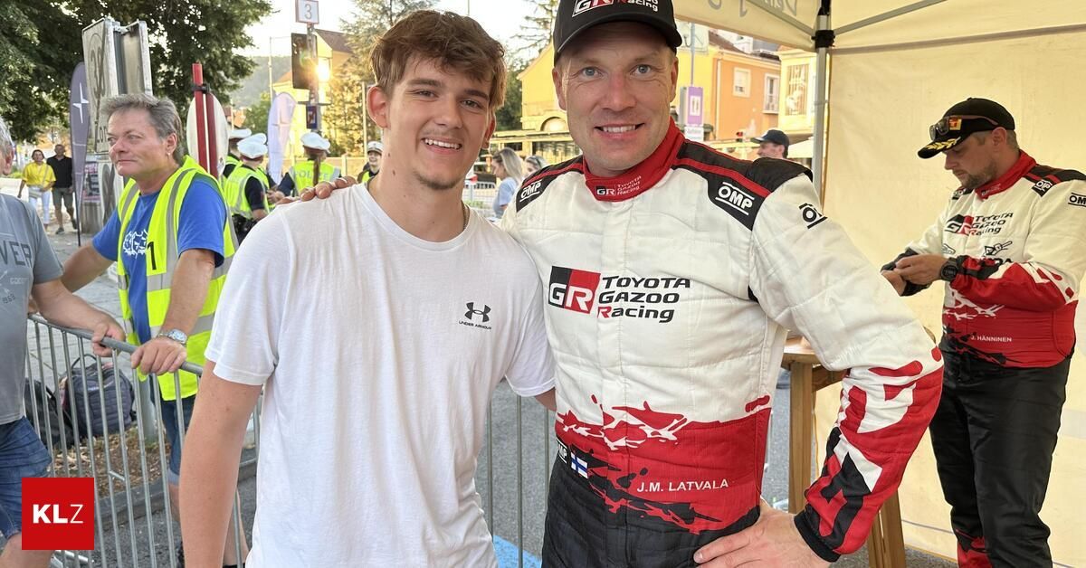 Two men pose for a photo in front of a tent, one wearing a white Under Armour shirt and the other in a red and white Toyota Gazoo Racing racing suit.
