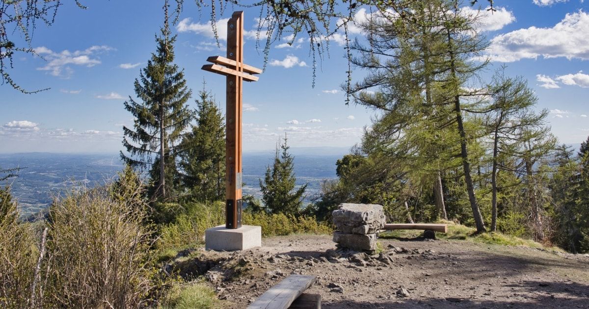 A wooden cross stands on a concrete base at a scenic overlook with a view of a valley and mountains.