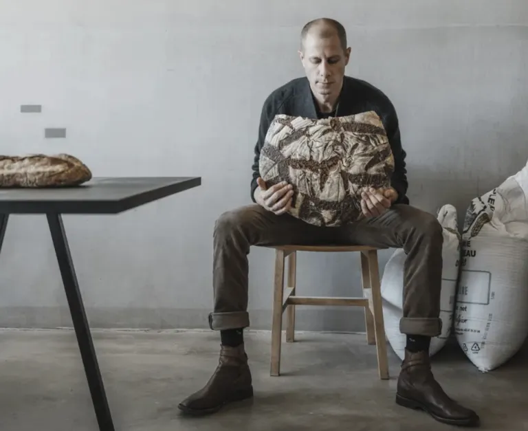 A man sits on a stool holding a large pillow shaped like a loaf of bread. He is in a room with a white wall and a table with a loaf of bread on it.