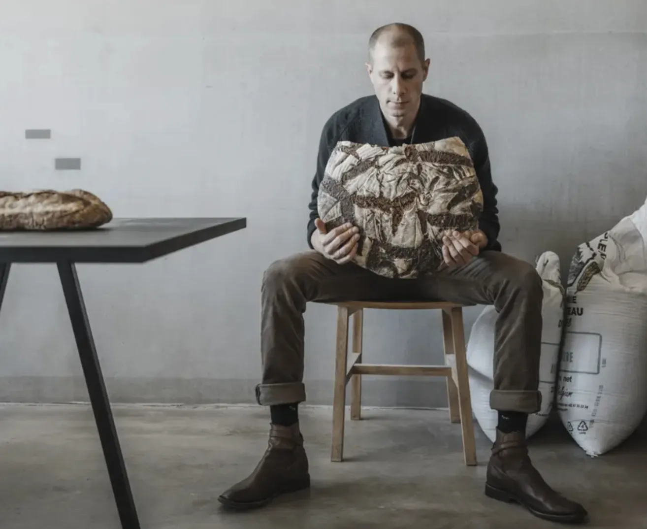 A man sits on a stool holding a large pillow shaped like a loaf of bread. He is in a room with a white wall and a table with a loaf of bread on it.