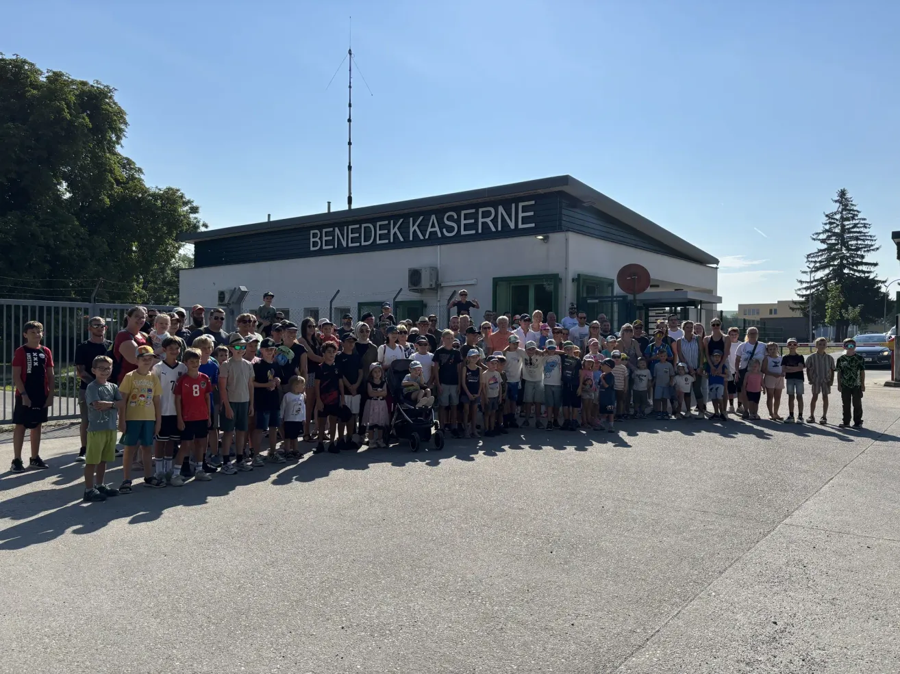 A large group of people, including children, are gathered in front of a building labeled 'Benedek Kaserne.' The building has a modern design with a flat roof and large windows. The sky is clear and blue.