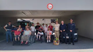 A group of people, some in wheelchairs, pose for a photo in a garage with a speed limit sign above. A basket of fruits is placed in front of them.