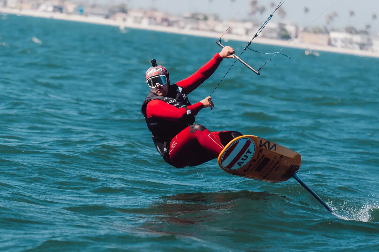 Ein Mann in einem roten Neoprenanzug und Helm windsurft auf dem Ozean, hält mit einer Hand die Stange und mit der anderen das Segel.