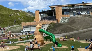 A large wooden structure resembling a snail with a green slide in a park with people playing and a modern building in the background.