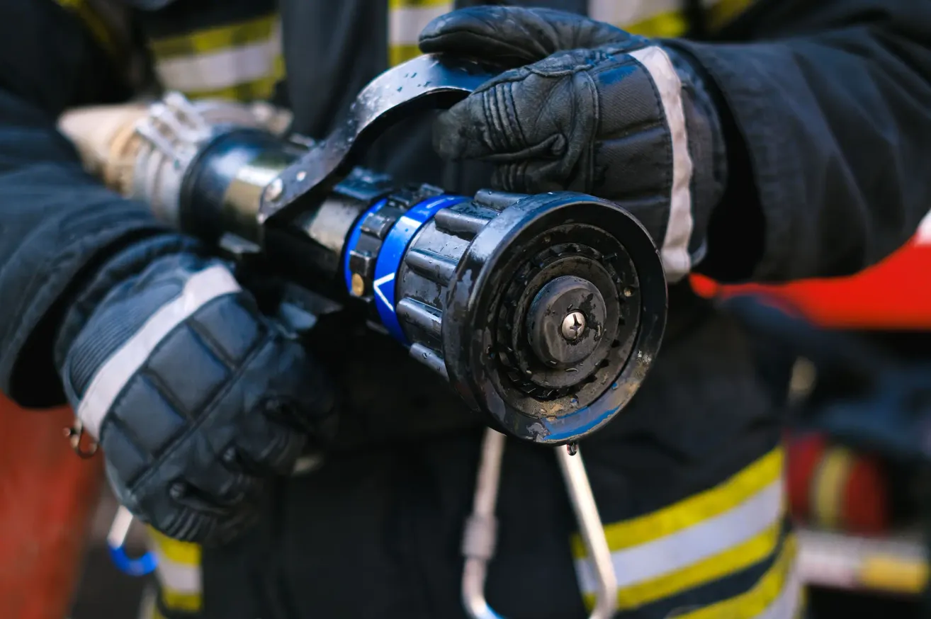 A firefighter in full gear holds a wet, blue-tipped fire hose nozzle close to the camera, with the nozzle's intricate design visible.