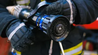 A firefighter in full gear holds a wet, blue-tipped fire hose nozzle close to the camera, with the nozzle's intricate design visible.