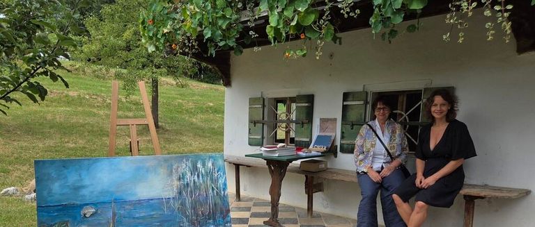 Two women sit on a bench outside a rustic house, one painting on a canvas, the other smiling. A table with books and a box is in front of them.