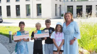 A group of children and an adult woman stand in front of a modern building, holding certificates. The children are smiling and appear to be posing for a photo.