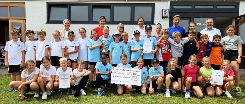 Eine Gruppe junger Fußballspieler, sowohl Jungen als auch Mädchen, posiert für ein Foto mit ihren Trainern und Eltern. Sie halten Urkunden und einen Scheck in der Hand, und einige tragen Mützen. Sie stehen vor einem Gebäude mit Glasfenstern und einem Schild, das 'Total' steht.