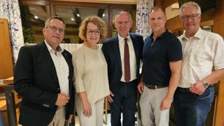 Four individuals, two men and two women, stand together smiling for a photo. The man on the left wears a black suit, the woman next to him wears glasses and a cream blouse. The man in the middle wears a dark suit and tie. The man on the right wears a navy blue polo shirt.