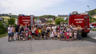 A group of adults and children pose for a photo in front of two fire trucks, with a clear sky and houses in the background.