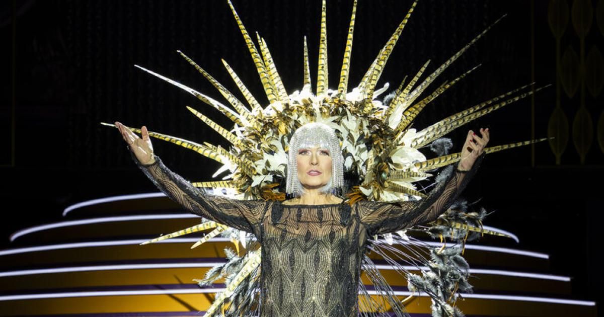 Eine Frau mit goldener Federkopfbedeckung und ausgestreckten Armen auf der Bühne.
