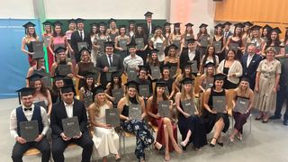 A group of graduates in academic attire, holding their diplomas, pose for a photo in a large room.