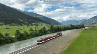 Ein roter Zug fährt entlang einer malerischen Route durch ein üppiges Tal, umgeben von Bergen und einem Fluss, unter einem teilweise bewölkten Himmel.