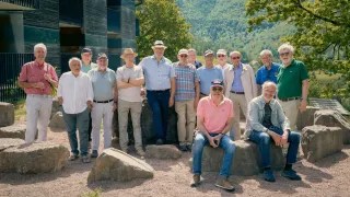 A group of older men dressed in casual attire, standing and sitting on rocks in front of a modern building with a mountainous backdrop.