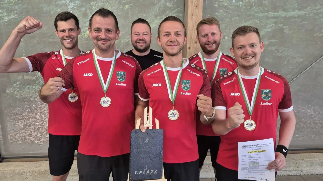 A group of men in red jerseys with medals around their necks are standing together and smiling. They are holding a plaque with the text '1. Platz'. The man in the middle is holding a trophy.