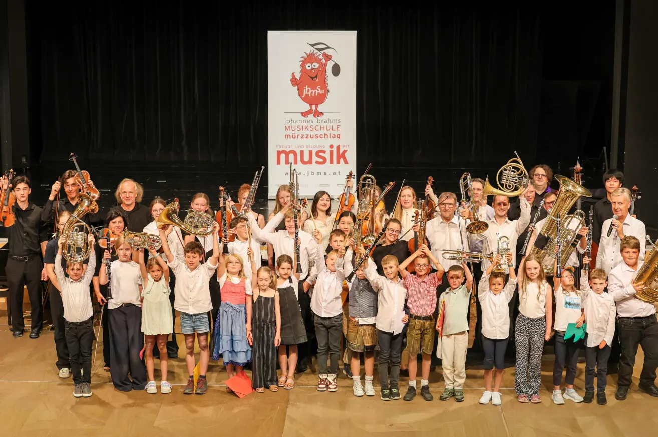 Eine Gruppe von Kindern und Erwachsenen posiert für ein Foto auf der Bühne und hält verschiedene Musikinstrumente in den Händen. Im Hintergrund befindet sich ein Banner mit dem Text 'Johannes Brahms Musikschule'.