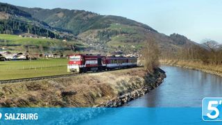 Ein roter Zug fährt auf einer Bahnstrecke in der Nähe eines Flusses, mit Bergen und einem Dorf im Hintergrund.