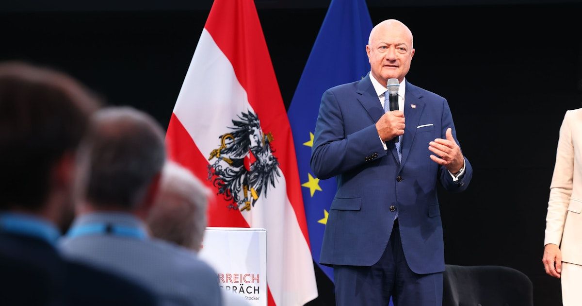 A man in a blue suit is giving a speech at a podium with flags behind him, one of which is the Austrian flag.