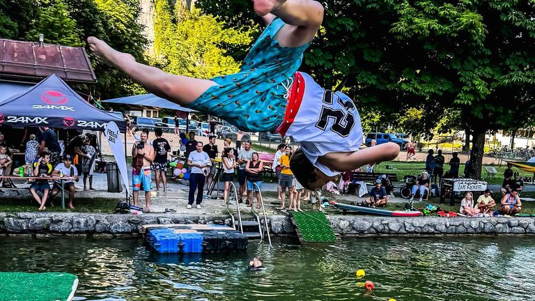 Ein junger Mensch taucht von einer Sprungbrett in einen Pool, trägt einen blauen Badeanzug mit rotem Gürtel und ein weißes Shirt mit der Nummer 23. Zuschauer beobachten von der Seite, und es gibt bunte Luftballons und ein Vordach im Hintergrund.