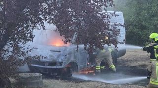 Firefighters extinguish a burning van near a tree with one firefighter using a hose and another standing by.