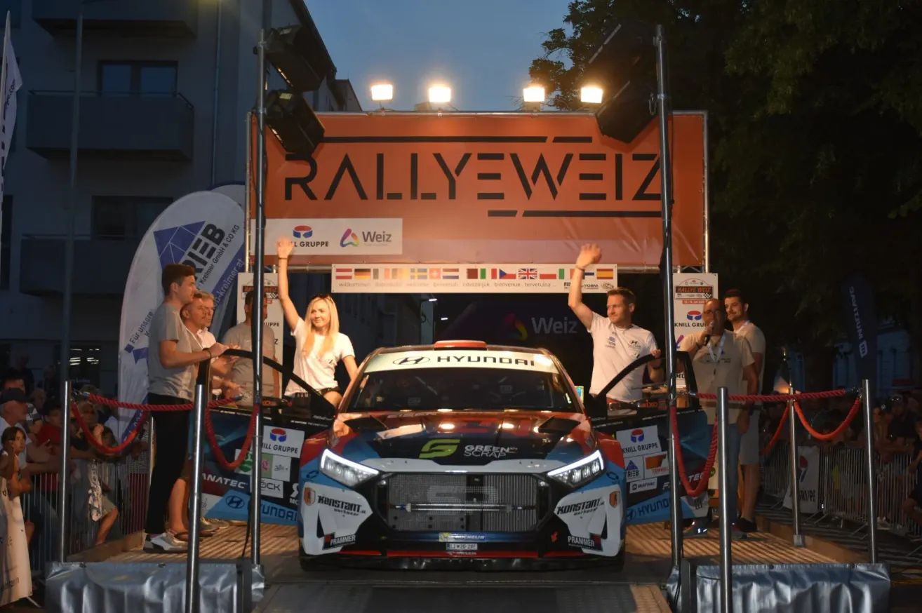 A rally car is on stage with two people standing in front of it. They are raising their hands and seem to be celebrating. The car has sponsors' logos on it. Behind the car, there is a banner with the text 'RALLYWEIZ' and some flags. There are people standing behind the car. There are lights on the stage.
