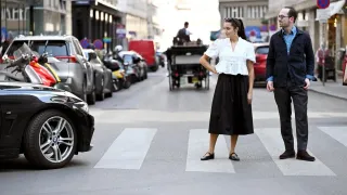 A woman in a white blouse and black skirt stands at a crosswalk, with a man in a dark suit beside her. Behind them, a horse-drawn carriage is moving on the road.