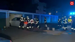 Firefighters in reflective gear are working on a white car in a parking lot at night. Two buckets are on the ground nearby.
