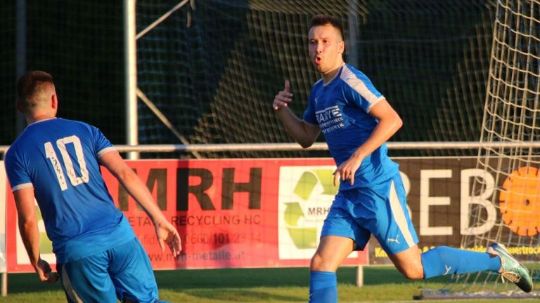 Ein Fußballspieler in blauer Uniform läuft mit konzentriertem Ausdruck auf das Tor zu. Hinter ihm ist ein weiterer Spieler teilweise sichtbar. Im Hintergrund befindet sich ein roter Banner mit Recycling-Informationen.