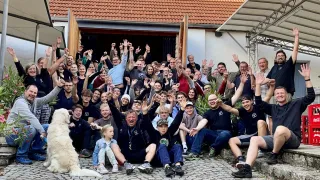 A large group of people, including adults and children, are gathered outside a building, smiling and waving. A dog is sitting on the ground nearby.