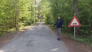 A man in a blue jacket is standing on a road with potholes, surrounded by dense green trees.