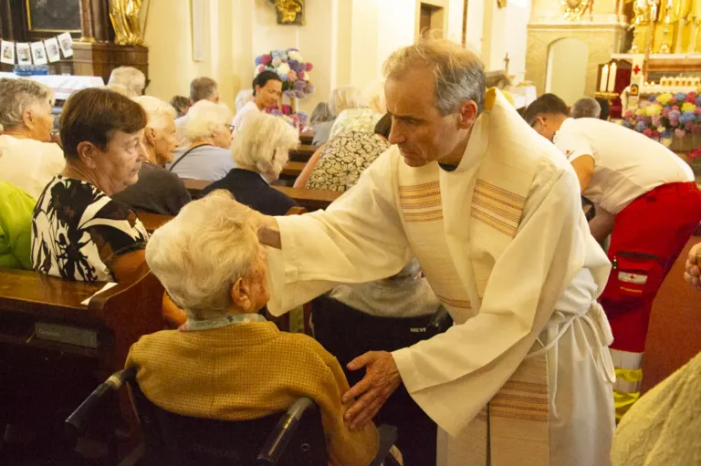 Ein Priester segnet eine ältere Frau im Rollstuhl in einer Kirche, umgeben von anderen älteren Personen.