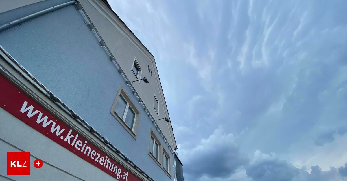 A building with a red sign that reads 'kleinezetung.at' on the side, with several windows and lamps attached to the wall under a cloudy sky.