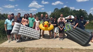 A group of people pose for a photo on a wooden deck, some holding inflatables, under a partly cloudy sky.