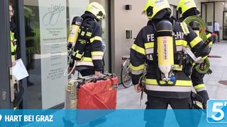 Firefighters in yellow and black uniforms with oxygen tanks on their backs are walking towards a building with a sign that reads 'Physiotherapie Institut'.