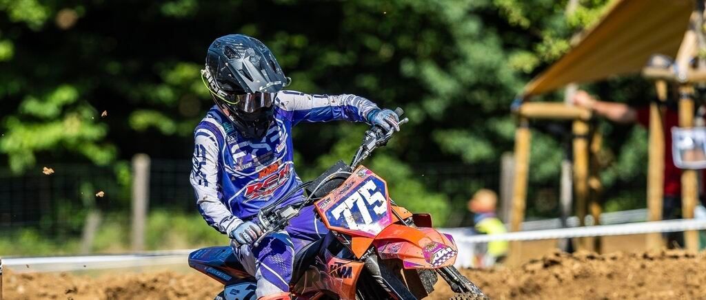 Ein Motocross-Fahrer in blauer und weißer Kleidung fährt ein orangefarbenes Motorrad mit der Nummer 775 auf einer Erdstrecke. Bäume und ein Schuppen sind im Hintergrund zu sehen.