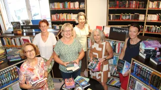 Five women stand in a library, smiling and holding books. They are surrounded by bookshelves filled with books. The shelves are labeled 'Biografie'.