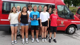 A group of young people, possibly students, are standing in front of a red vehicle, smiling and posing for a photo. The boy in the middle holds a plaque.