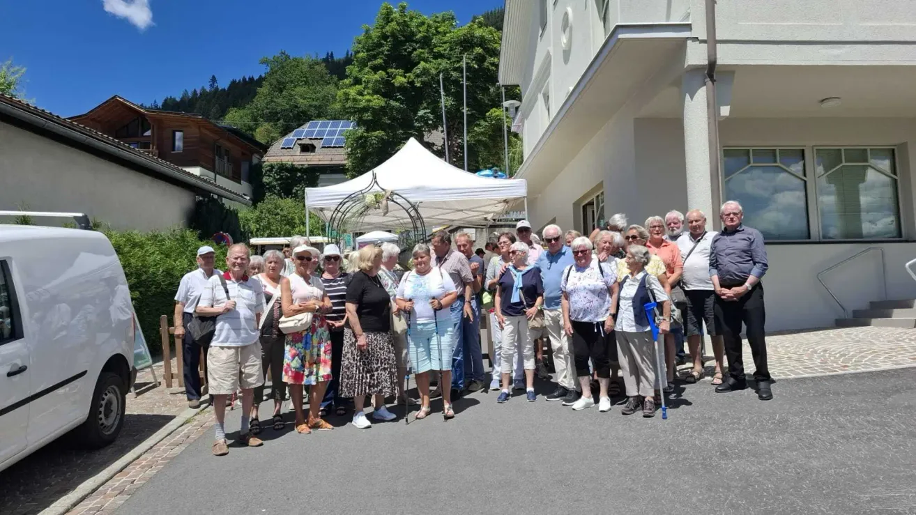 A group of elderly people are standing in front of a building, some wearing hats and glasses, with a white tent in the background.