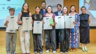 Five women and three men stand on a stage, holding certificates. The women wear casual clothing, and the men wear suits. They all smile for the camera.