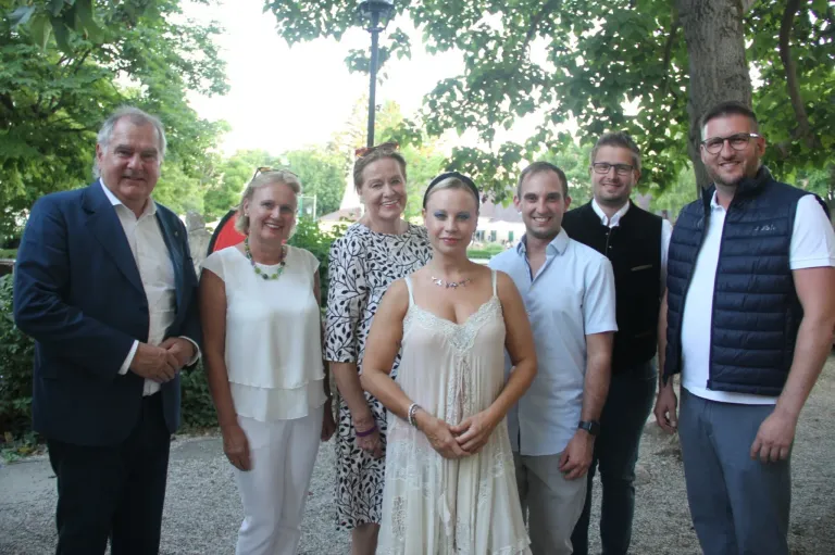 A group of people dressed in summer attire are posing for a photo in a park with trees and a lamp post in the background.
