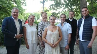 A group of people dressed in summer attire are posing for a photo in a park with trees and a lamp post in the background.