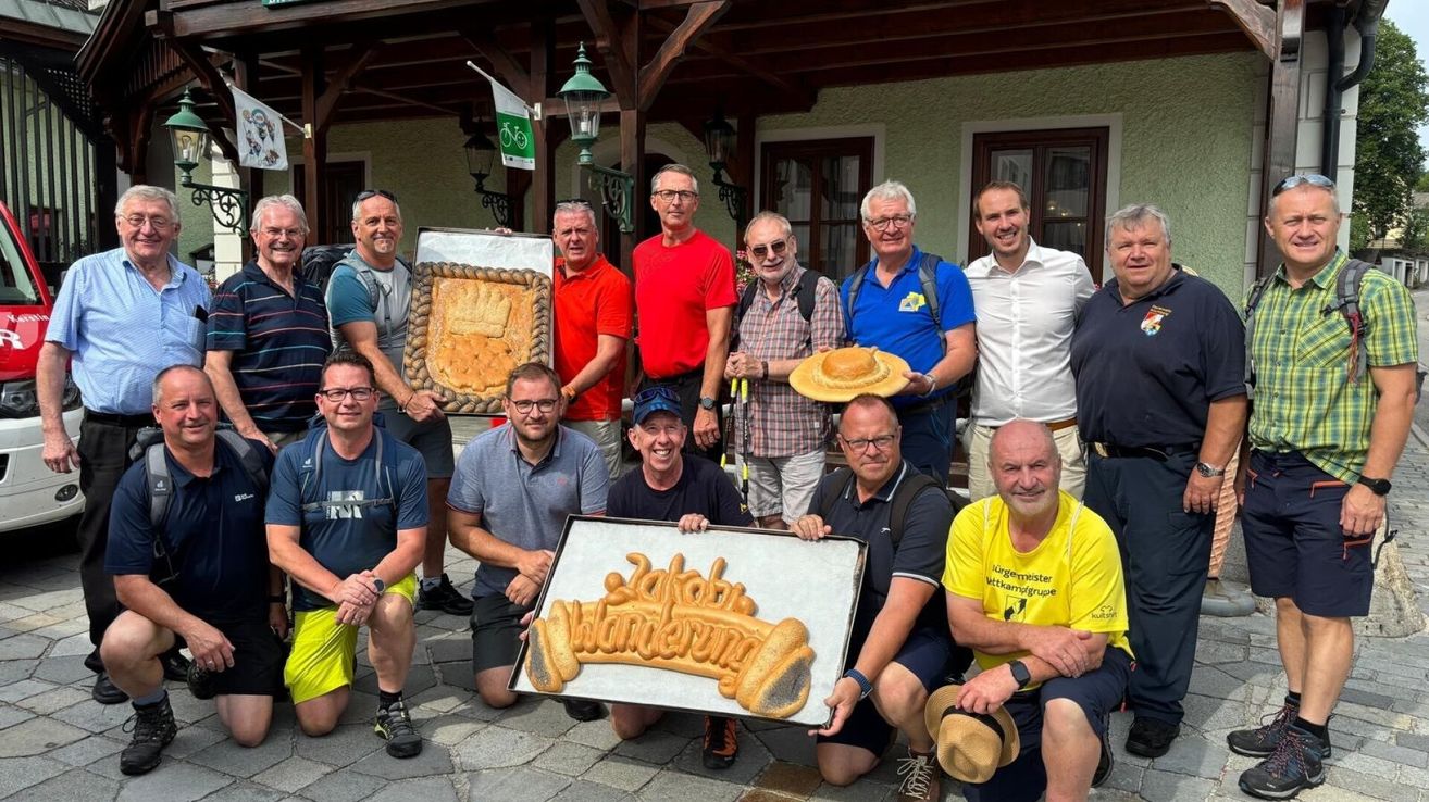 Eine Gruppe von Männern in Freizeitkleidung, einige mit Hüten, posiert für ein Foto vor einem Gebäude mit Fahrradfahne und Lampe. Ein Mann hält ein großes Brot mit Kronendesign, während ein anderer eine Tablett mit einer Brotskulptur hält.
