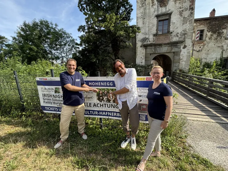 Drei Personen stehen vor einem Schild, das eine kulturelle Veranstaltung ankündigt, lächeln und posieren für ein Foto. Das Schild trägt den Namen 'Hohenegg' und enthält Datum und Uhrzeit der Veranstaltung.