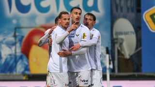 Four soccer players in white uniforms celebrate a goal on a rainy field. The player in the center points to the sky.