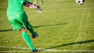A soccer player in green uniform is jumping to catch the ball. The ball is in the air, and another player is in the background.