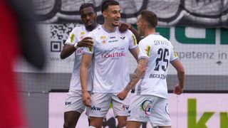 Three soccer players in white uniforms celebrate a goal on the field. The player in the middle wears a white shirt with the word 'Dortmund' and the number 25 on the back. The player on the left wears a white shirt with the word 'DC' on it. The player on the right has a tattoo on his arm.