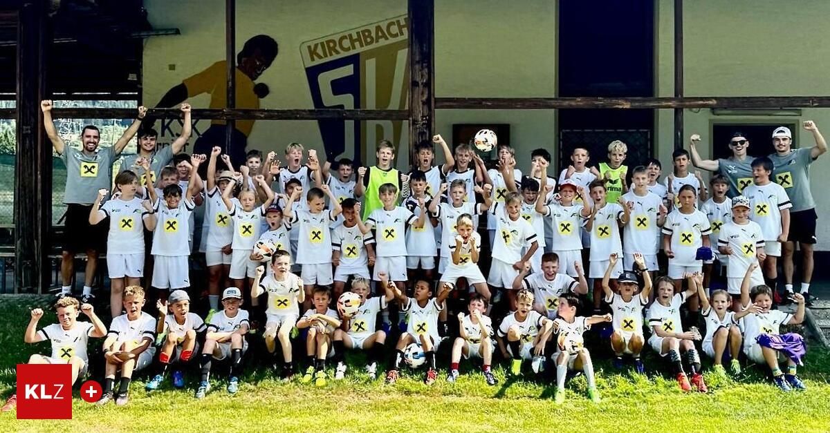 A group of young soccer players in white uniforms with yellow X's pose for a photo on a grassy field. Behind them is a wall with a large soccer player's image and the word Kirchbach.