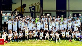 A group of young soccer players in white uniforms with yellow X's pose for a photo on a grassy field. Behind them is a wall with a large soccer player's image and the word Kirchbach.